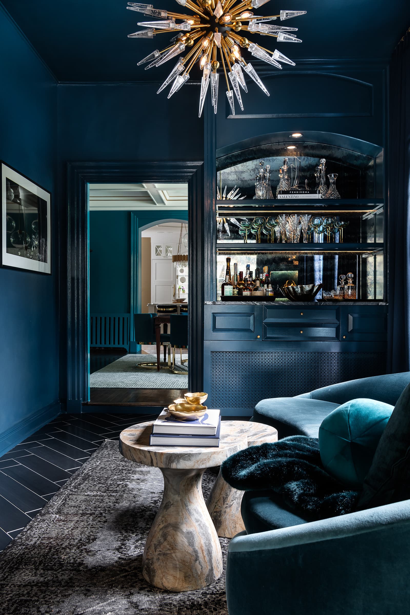 Moody lounge with dark blue walls, built-in bar, velvet seating, and statement chandelier in a dramatic residential interior
